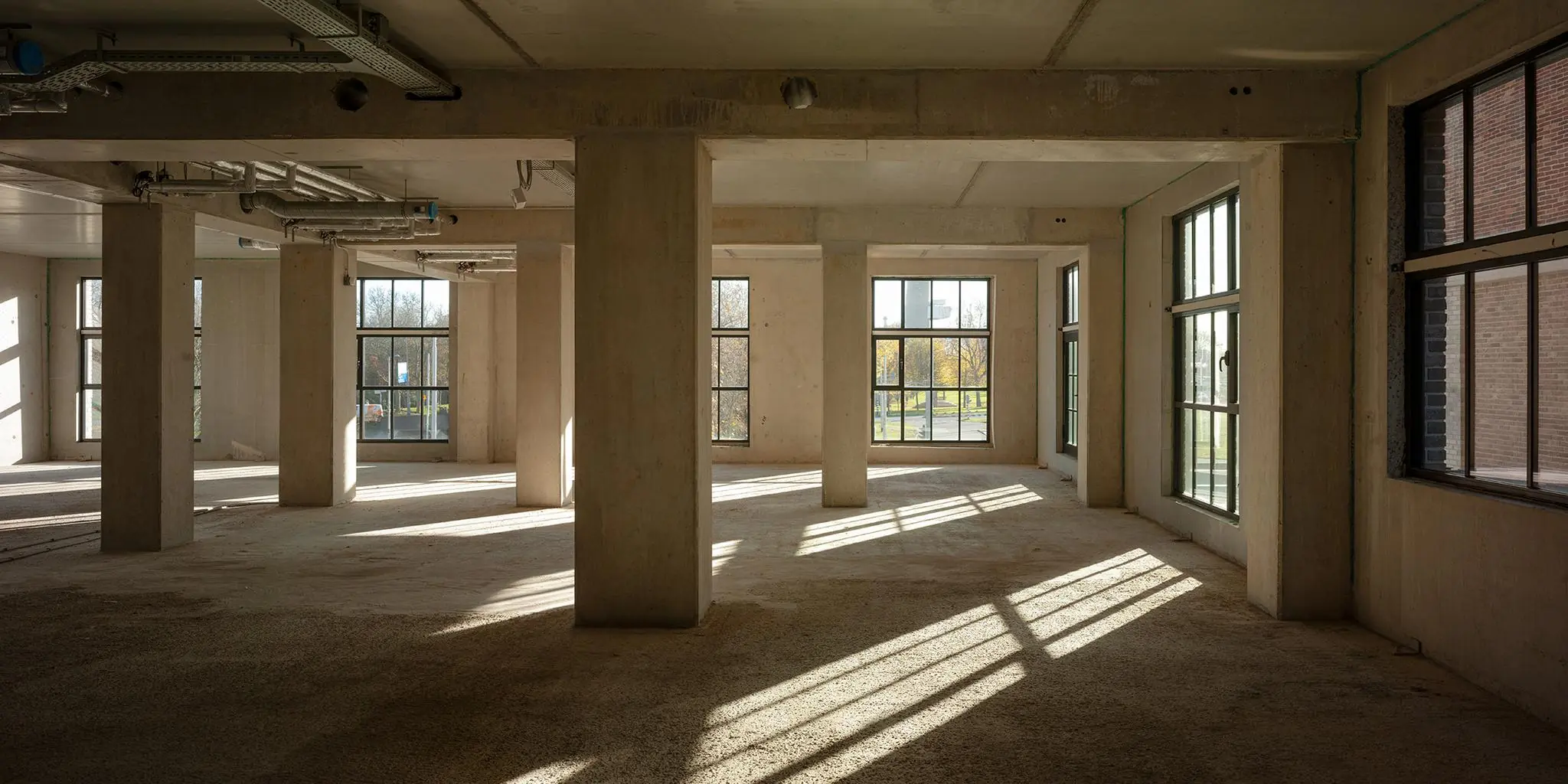 Leegstaand betonnen interieur met grote ramen aan de Karl Weisbardstraat, waar zonlicht strepen op de vloer werpt.